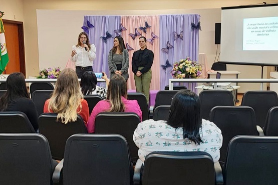 A imagem mostra uma apresentação em um auditório. Três mulheres estão em pé na frente da sala, de frente para o público. Elas estão em um palco decorado com cortinas lilás e rosas e borboletas roxas. Há arranjos de flores ao lado delas. O público está sentado em cadeiras pretas, e a maioria das pessoas está de costas para a câmera. À direita, há uma tela de projeção com um slide que parece ter texto, mas não é legível na imagem. No canto esquerdo, há uma bandeira. A atmosfera parece ser de um evento formal ou uma palestra.