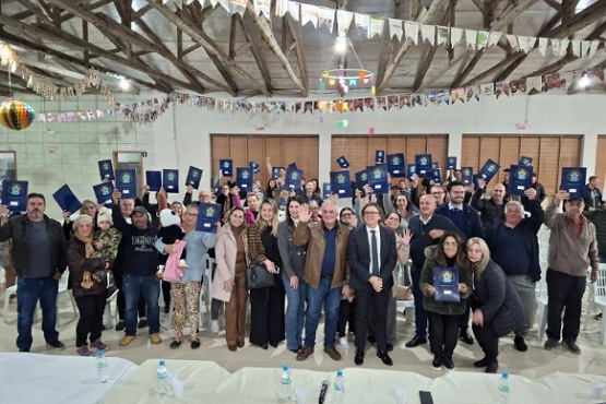 A imagem mostra um grupo grande de pessoas posando para uma foto em um ambiente interno. A maioria das pessoas está segurando um documento ou certificado azul, levantado acima de suas cabeças. O local parece ser um salão com teto de madeira exposta e bandeirinhas coloridas penduradas, sugerindo uma decoração típica de festa junina. Na frente do grupo, há uma mesa com garrafas de água e alguns papéis. As pessoas estão sorrindo e parecem estar celebrando uma ocasião especial.