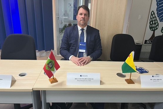 A imagem mostra um homem de terno escuro e gravata sentado em uma mesa de conferência. Ele tem cabelo castanho escuro e barba. Na mesa à sua frente, há uma placa com o nome "Dr.  Luiz Felipe Schuch" e o título "Desembargador do TJSC". À esquerda da placa, há uma pequena bandeira do estado de Santa Catarina, e à direita, uma bandeira do estado do Acre. Ao fundo, há uma parede de vidro e cortinas azuis. A sala parece ser um ambiente de reunião ou conferência.