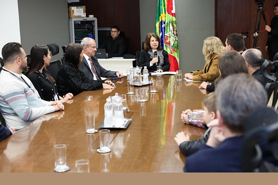 A imagem mostra uma reunião formal em uma sala de conferências. Há várias pessoas sentadas ao redor de uma mesa de madeira retangular. No centro da mesa, há garrafas de água e copos. Uma mulher, que parece estar liderando a reunião, está falando ao microfone. Ela está sentada na ponta da mesa. Ao fundo, há bandeiras do Brasil e de Santa Catarina, e um homem em pé, que parece estar operando equipamentos de áudio ou vídeo. As pessoas estão vestidas de maneira formal, sugerindo que se trata de um evento oficial ou governamental.