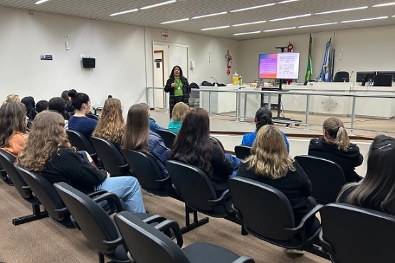 A imagem mostra uma sala de conferências ou auditório com várias pessoas sentadas em cadeiras pretas, voltadas para a frente da sala. Na frente, há uma mulher de pé, aparentemente dando uma apresentação. Ela está ao lado de uma tela que exibe um slide com texto, mas o conteúdo não é legível na imagem. No fundo, há uma bancada com microfones e bandeiras, sugerindo que o local pode ser uma sala de reuniões oficial ou governamental. As paredes são claras e o teto tem iluminação embutida. A maioria das pessoas na audiência parece ser do sexo feminino.