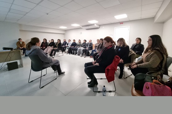 A imagem mostra uma sala de reunião com várias pessoas sentadas em cadeiras dispostas em um semicírculo. A sala tem um teto com iluminação embutida e paredes claras. No centro do semicírculo, uma mulher está sentada de frente para o grupo, aparentemente conduzindo a reunião ou palestra. As pessoas estão vestidas de forma casual, com roupas de inverno, como casacos e cachecois. Há uma mesa pequena com uma garrafa de água no chão, próxima ao grupo. A atmosfera parece ser de atenção e concentração.