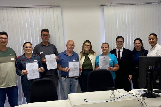 A imagem mostra um grupo de nove pessoas posando para uma foto em um ambiente de escritório. Quatro delas estão segurando certificados ou documentos em suas mãos. As pessoas estão alinhadas em pé, lado a lado, e parecem estar participando de algum tipo de cerimônia ou evento de entrega de certificados. Ao fundo, há persianas brancas cobrindo as janelas. Na frente do grupo, há uma mesa com um monitor de computador e alguns cabos. Todos estão sorrindo e vestidos de maneira casual ou formal.