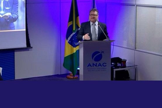 A imagem mostra um homem de terno e gravata falando em um púlpito. Atrás dele, há uma bandeira do Brasil e um painel com iluminação azul. No púlpito, está o logotipo da ANAC, que é a Agência Nacional de Aviação Civil do Brasil. À esquerda, há uma tela de projeção com uma imagem parcialmente visível de uma pessoa segurando algo nas mãos. O ambiente parece ser uma sala de conferências ou um auditório.