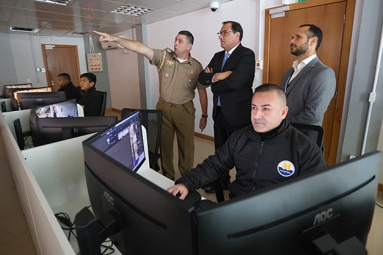 A imagem mostra uma sala de controle com várias pessoas. No primeiro plano, há um homem sentado em frente a um computador, usando um uniforme preto com um emblema no peito. Ele está olhando para a tela do computador. Ao fundo, há mais três homens em pé. Um deles, que parece ser um oficial militar, está apontando para algo fora da imagem. Ele está ao lado de dois homens de terno, que observam na mesma direção. No lado esquerdo da imagem, há mais duas pessoas sentadas em frente a computadores, também concentradas nas telas. A sala tem um ambiente moderno, com várias telas de computador e um teto com iluminação embutida.