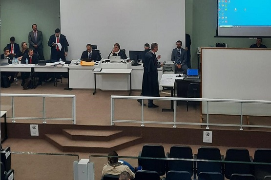 A imagem mostra uma sala de tribunal. Há várias pessoas sentadas em uma longa mesa em formato de "L", que parece ser a bancada dos advogados ou juízes. Algumas pessoas estão de pé, enquanto outras estão sentadas, aparentemente ouvindo ou discutindo algo. Um homem de terno e toga está em pé no centro da sala, possivelmente apresentando um argumento ou falando com o juiz. No fundo, há uma grande tela de projeção com um computador ligado, exibindo a área de trabalho do Windows. A sala tem várias fileiras de cadeiras vazias na parte inferior da imagem, sugerindo que é um espaço para o público ou para outras partes envolvidas no julgamento. A atmosfera é formal e típica de um ambiente jurídico.