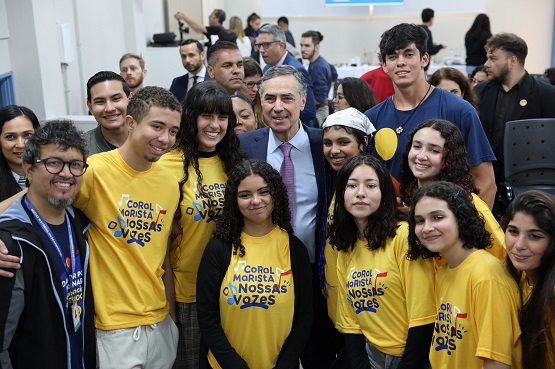 A imagem mostra um grupo de pessoas posando para uma foto. No centro, há um homem de terno e gravata, que parece ser uma figura pública ou autoridade. Ao redor dele, há várias pessoas, principalmente jovens, muitos dos quais estão vestindo camisetas amarelas com a inscrição "Coral Marista Nossas Vozes". Todos parecem estar sorrindo e felizes. Ao fundo, há outras pessoas e um ambiente que parece ser um espaço interno, possivelmente um auditório ou sala de eventos.