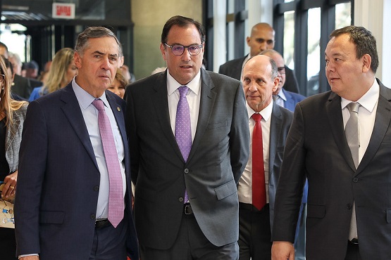 A imagem mostra três homens caminhando juntos em um ambiente interno, possivelmente um corredor de um prédio. Todos estão vestidos formalmente com ternos. O homem à esquerda está usando um terno azul escuro com uma gravata lilás clara. Ao lado dele, o segundo homem veste um terno cinza escuro com uma gravata lilás. O terceiro homem, à direita, veste um terno cinza escuro com uma gravata branca. Ao fundo, há outras pessoas e uma parede de vidro que permite a entrada de luz natural. A cena parece ser de um evento formal ou reunião.