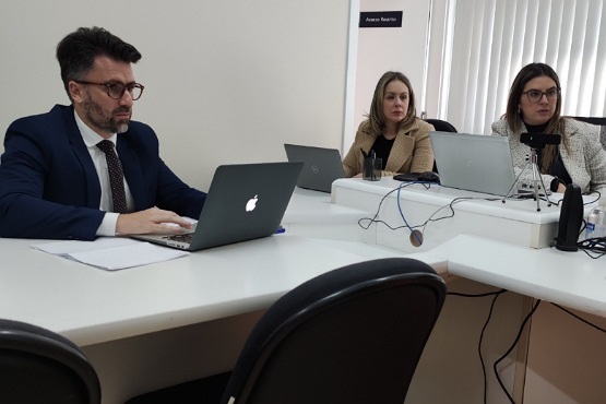 A imagem mostra três pessoas sentadas em uma mesa de júri no formato em T. À esquerda, um homem de terno azul escuro, camisa branca e gravata preta está usando um laptop da Apple. Ele tem cabelo escuro e barba. À direita, na ponta da mesa, duas mulheres estão sentadas lado a lado. Ambas estão usando laptops e parecem estar prestando atenção em algo à frente. A mulher do meio tem cabelo loiro e veste um casaco bege. A mulher à direita tem cabelo castanho e usa óculos, além de um casaco claro. Na mesa, há também uma webcam e alguns cabos. Ao fundo, há uma parede clara com uma placa que diz "Acesso Restrito" e uma janela com persianas verticais.
