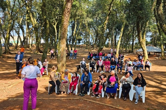 A imagem mostra um grupo de pessoas reunidas em um ambiente ao ar livre, em uma área arborizada. Há várias árvores ao redor e o chão é de terra. As pessoas estão sentadas em cadeiras de plástico branco, dispostas em fileiras. A maioria delas parece ser de mulheres, e algumas estão conversando entre si. No canto esquerdo da imagem, uma mulher de costas, vestindo calça roxa e camiseta branca, parece estar falando ou apresentando algo para o grupo. O clima parece ser de um encontro social ou uma atividade comunitária em um parque. A luz do sol atravessa as árvores, criando sombras no chão.
