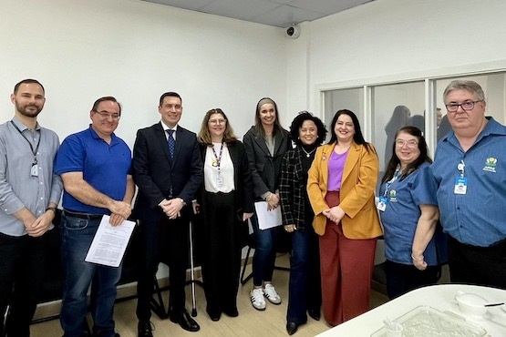 A imagem mostra um grupo de nove pessoas posando para uma foto em um ambiente interno, que parece ser uma sala de reuniões. As pessoas estão alinhadas lado a lado, sorrindo para a câmera. Da esquerda para a direita, o primeiro homem está usando uma camisa azul clara e calça escura. O segundo homem veste uma camisa polo azul escura e segura um papel. O terceiro homem está de terno escuro e gravata, com as mãos cruzadas à frente. A primeira mulher está usando um casaco preto sobre uma blusa branca e calça preta, com um colar. A segunda mulher veste um casaco cinza e calça jeans, segurando um papel. A terceira mulher está com um casaco xadrez preto e cinza, e calça preta. A quarta mulher usa um casaco mostarda sobre uma blusa lilás e calça vermelha. A quinta mulher está com uma camisa azul e crachá, ao lado de um homem que também veste uma camisa azul com crachá. O ambiente tem paredes brancas e janelas de vidro ao fundo. Na frente, há uma mesa branca com alguns objetos em cima.