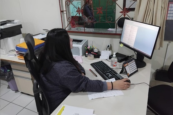 A imagem mostra uma mulher sentada em uma cadeira de escritório, trabalhando em frente a um computador. Ela está usando um moletom cinza escuro e tem cabelos longos e escuros. A mesa está organizada com um teclado, um mouse, um monitor grande, um frasco de álcool em gel e alguns papéis. Ao fundo, há uma janela com grades decorativas e uma cortina bege. Do lado esquerdo, há uma impressora e uma pilha de pastas amarelas. A sala parece ser um escritório, com piso de azulejos brancos.