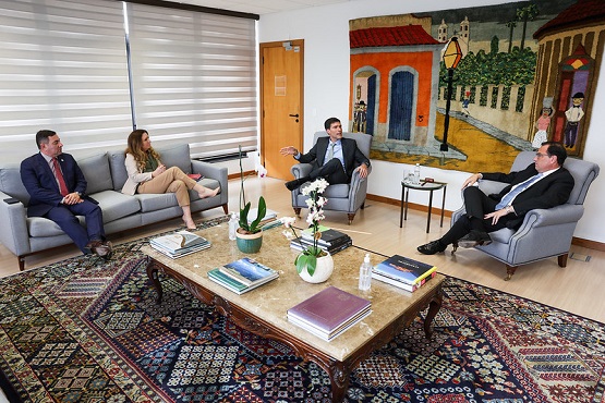 A imagem mostra uma sala de estar ou de reunião com quatro pessoas sentadas em sofás. A sala tem um tapete colorido no chão e uma mesa de centro com alguns objetos sobre ela, incluindo livros e plantas. Na parede ao fundo, há uma grande pintura colorida. As pessoas estão vestidas formalmente, com ternos e roupas sociais.