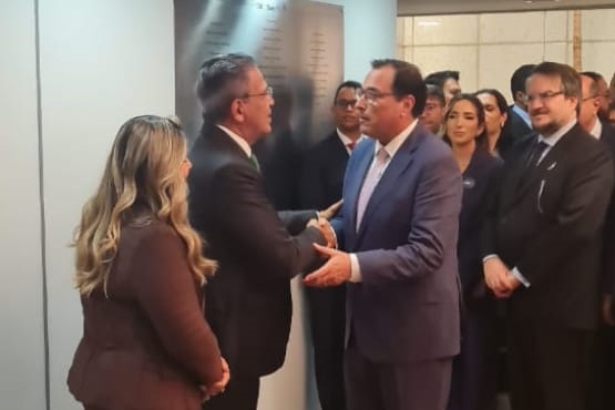 A imagem mostra um grupo de pessoas em um ambiente interno, possivelmente em um evento formal ou reunião. No centro, há um homem de terno azul claro e óculos, que parece estar falando ou gesticulando para uma mulher de cabelos loiros e longos, vestida com um terno marrom. Ao fundo, há várias outras pessoas, a maioria delas usando trajes formais, como ternos escuros.