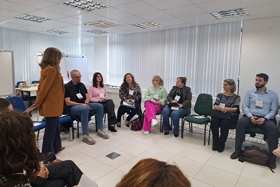 A imagem mostra uma sala de conferência onde várias pessoas estão sentadas em cadeiras dispostas em um círculo. As cadeiras são de cor azul e verde. As pessoas parecem estar participando de uma reunião ou workshop, algumas estão fazendo anotações. No fundo da sala, há uma tela de projeção branca. A sala é bem iluminada, com luzes fluorescentes no teto.