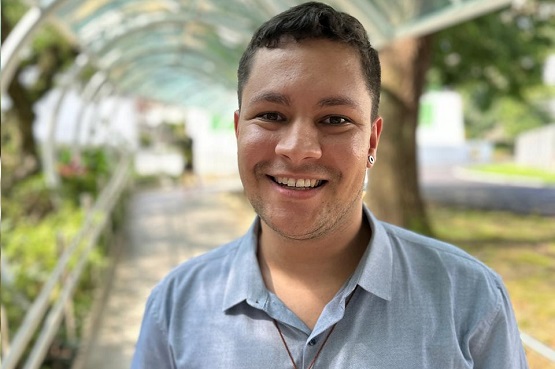 A imagem mostra um homem jovem sorrindo para a câmera. Ele tem pele clara, cabelo curto e escuro, e está usando uma camisa azul clara. Ele também tem um brinco na orelha esquerda. O fundo da imagem é um ambiente ao ar livre, com uma estrutura de arco transparente acima dele e árvores ao redor, sugerindo que ele está em um caminho ou passarela coberta. A iluminação é clara e parece ser um dia ensolarado.