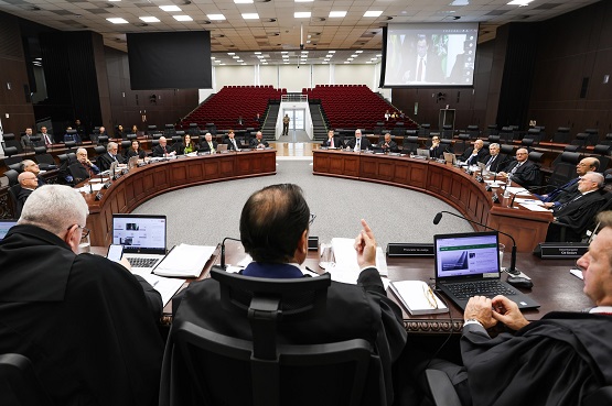 A imagem mostra uma sala de tribunal. Há uma mesa em formato de U, onde várias pessoas estão sentadas, todas vestidas com trajes formais, possivelmente togas, sugerindo que são magistrados. No centro da imagem, de costas para a câmera, está uma pessoa que parece estar presidindo a sessão. Na frente dela, há laptops abertos e documentos. Ao fundo, há uma fileira de cadeiras vermelhas vazias e uma tela de projeção que exibe uma imagem. A sala é bem iluminada e tem um teto com várias luzes.