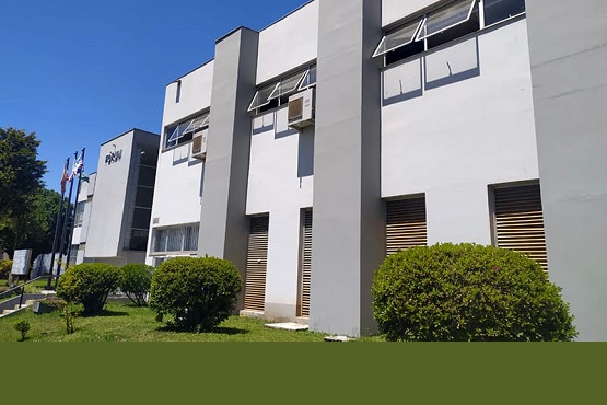 A imagem mostra um edifício de dois andares com uma fachada moderna e de cor clara. Há várias janelas com persianas e algumas delas estão abertas. No lado esquerdo da imagem, há bandeiras hasteadas, sugerindo que o local pode ser uma instituição pública ou uma empresa. Em frente ao prédio, há um gramado bem cuidado com arbustos arredondados. O céu está azul, indicando que é um dia ensolarado.