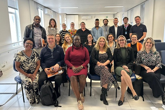 A imagem mostra um grupo de 21 pessoas posando para uma foto em uma sala. Elas estão organizadas em três fileiras: as duas fileiras da frente estão sentadas em cadeiras, enquanto a fileira de trás está em pé. A maioria das pessoas está sorrindo. A sala parece ser um ambiente de escritório ou sala de conferências, com paredes brancas e janelas ao fundo. As pessoas estão vestidas de maneira casual e formal, com uma variedade de estilos e cores de roupas. A atmosfera parece ser amigável e profissional.