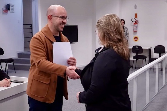 A imagem mostra duas pessoas em um ambiente interno. À esquerda, um homem careca e de óculos, vestindo um casaco marrom e segurando um papel, está apertando a mão de uma mulher à direita. A mulher tem cabelos loiros e usa um casaco preto. Ambos parecem estar sorrindo, sugerindo que é um momento de congratulação ou entrega de certificado. Ao fundo, há algumas cadeiras vazias, uma escada e uma pessoa observando a cena. Há também um extintor de incêndio na parede.