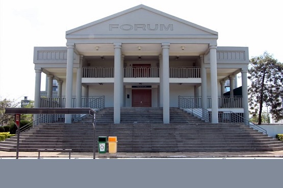 A imagem mostra a fachada de um edifício com arquitetura clássica, identificado como "FÓRUM" na parte superior. O prédio tem um estilo que lembra templos gregos, com colunas na frente e um frontão triangular no topo. Há uma escadaria larga que leva até a entrada principal, que possui uma porta de madeira. Em frente à escadaria, há lixeiras de coleta seletiva, uma verde e outra amarela, e um ponto de ônibus. O céu está claro e há algumas árvores ao fundo, sugerindo que o local está em uma área arborizada.