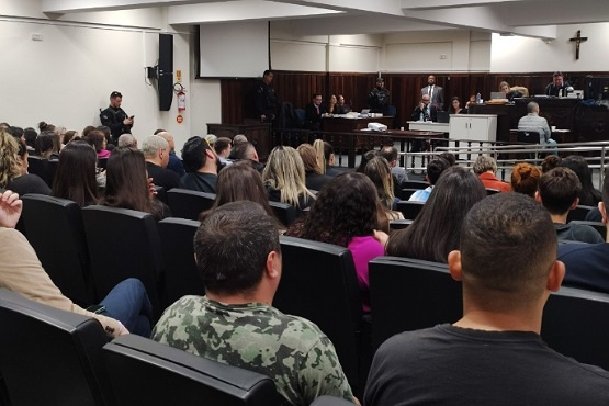 A imagem mostra uma sala de tribunal com várias pessoas sentadas em bancos, assistindo a um julgamento. A sala está cheia, com pessoas de diferentes idades e estilos de cabelo. Na frente, há uma mesa onde estão sentados os juízes ou advogados, e uma pessoa está em pé, possivelmente falando ou apresentando algo. Na parede, há um crucifixo.
