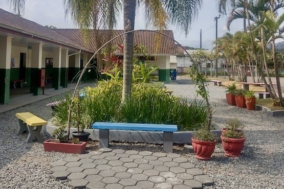A imagem mostra um pátio ao ar livre, provavelmente de uma escola ou instituição similar. No centro, há um banco de concreto pintado de azul, cercado por vasos de plantas. O chão é coberto por pedras pequenas e há uma área pavimentada com blocos hexagonais em frente ao banco. Atrás do banco, há um canteiro elevado com plantas e uma palmeira. À esquerda, há um prédio com um corredor coberto, com paredes pintadas de branco e verde escuro. À direita, há mais vasos de plantas e algumas mesas de piquenique. O ambiente é bem cuidado e tem várias palmeiras ao redor.