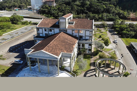A imagem mostra uma vista aérea de um edifício identificado como "Fórum". O prédio tem um design clássico com colunas na entrada e um telhado de telhas marrons. Ao lado do edifício, há um estacionamento com algumas vagas ocupadas por carros. A área ao redor é arborizada, com muitas árvores e vegetação. Ao fundo, é possível ver uma estrada e mais vegetação, sugerindo que o local está em uma área com bastante natureza ao redor. Há também uma estrutura circular aberta, semelhante a um gazebo, próxima ao edifício principal.