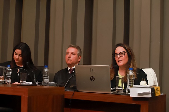 A imagem mostra três pessoas sentadas atrás de uma mesa de madeira em um ambiente que parece ser uma sala de conferências ou um tribunal. À esquerda, uma mulher de cabelos longos e escuros está olhando para baixo, possivelmente lendo algo. No centro, um homem de terno e gravata está olhando para frente com uma expressão neutra. À direita, uma mulher de óculos está falando, com um laptop HP aberto à sua frente. Na mesa, há garrafas de água, copos e papeis. Ao fundo, há uma parede com paineis ripados na madeira escura.
