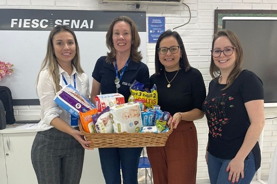 A imagem mostra quatro mulheres sorrindo e posando para a foto em um ambiente interno. Elas estão segurando juntas uma cesta cheia de produtos, que parecem ser itens de higiene pessoal e alimentos, como fraldas, lenços umedecidos e pasta de dentes. Ao fundo, há uma parede branca com um letreiro que diz "FIESC SENAI". As mulheres estão vestidas de maneira casual, com blusas e calças. O ambiente parece ser uma sala de aula ou um espaço de reunião, com móveis brancos e um quadro na parede.