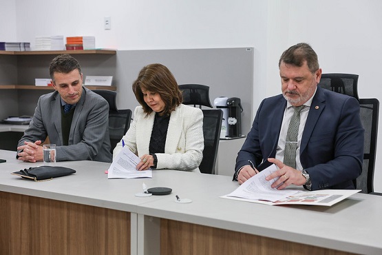 A imagem mostra três pessoas sentadas lado a lado em uma mesa de reunião. À esquerda, há um homem de terno cinza escuro, camisa azul escura e gravata azul também, olhando para a assinatura da mulher ao seu lado esquerdo. No centro, uma mulher de cabelo castanho e liso, vestindo um blazer branco sobre uma blusa preta, está assinando um documento. À direita, um homem de terno azul escuro, camisa branca e gravata cinza com padrão, também está assinando um documento. A mesa é de madeira clara e há alguns objetos sobre ela, como um copo de água, uma carteira e papéis. Ao fundo, há uma estante com livros e uma cafeteira. A cena parece ser de uma reunião formal ou assinatura de contrato.