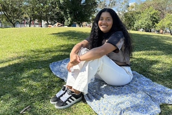 A imagem mostra uma mulher sentada em um gramado, sobre um tecido azul com estampas. Ela está sorrindo e olhando para a câmera. A mulher tem cabelos longos e cacheados, veste uma camiseta de manga curta em tons escuros e calças brancas. Nos pés, ela usa tênis pretos com detalhes brancos. Ao fundo, há árvores e um céu azul claro, sugerindo que é um dia ensolarado em um parque.
