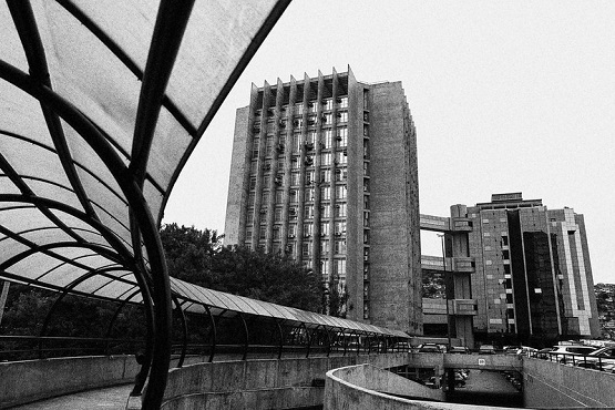 A imagem em preto e branco mostra uma cena urbana com dois edifícios altos e modernos. O edifício à esquerda é mais alto, com uma fachada de concreto e várias janelas verticais. O edifício à direita é um pouco mais baixo e tem um design semelhante, com uma estrutura de concreto e janelas. No primeiro plano, há uma passarela coberta com uma estrutura curva e transparente, que parece ser feita de vidro ou plástico, sustentada por armações metálicas. A passarela segue em direção aos edifícios, criando uma linha curva que guia o olhar para o fundo da imagem. Há também algumas árvores visíveis ao lado dos edifícios. O céu está claro, mas a imagem não mostra muitos detalhes devido ao efeito monocromático.