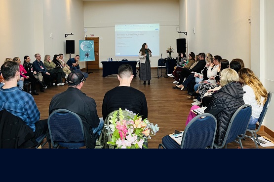 A imagem mostra uma sala de conferências com um grupo de pessoas sentadas em cadeiras dispostas em um semicírculo. No centro, uma mulher está de pé, aparentemente fazendo uma apresentação ou discurso. Ao fundo, há uma tela de projeção com um slide exibido, mas o texto não está legível na imagem. As paredes são claras e o piso é de madeira. Há também um banner ao lado da tela e algumas flores em um vaso no chão. As pessoas na sala parecem estar prestando atenção à apresentação.