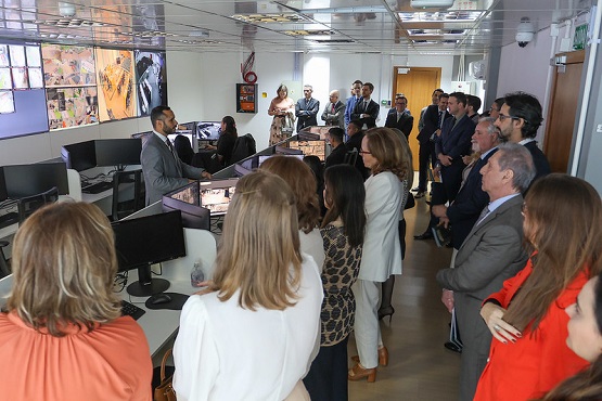 A imagem mostra uma sala de controle ou centro de operações com várias pessoas reunidas. Há um grupo de cerca de trinta pessoas, a maioria vestida formalmente, como em um ambiente de trabalho ou evento corporativo. Elas estão de pé, algumas olhando para um homem que parece estar apresentando ou explicando algo. Ele está de pé ao lado de uma mesa com vários monitores de computador. Nas paredes, há telas grandes exibindo imagens ou vídeos, possivelmente de câmeras de segurança ou monitoramento. A sala é bem iluminada e tem um ambiente moderno e profissional.
