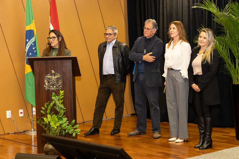 A imagem mostra cinco pessoas em um ambiente formal. À esquerda, uma mulher está em pé atrás de um púlpito de madeira, falando ou apresentando algo. Ela usa óculos e tem cabelos longos e escuros. Ao lado dela, há quatro pessoas em pé, lado a lado. O primeiro homem à esquerda veste uma jaqueta de couro preta, camisa azul clara e calças marrons. O segundo homem usa um terno azul escuro e está com os braços cruzados. Ao lado dele, uma mulher veste uma blusa branca e calças cinzas, e a última mulher à direita usa uma blusa clara e um casaco preto, com botas pretas. No fundo, há duas bandeiras, uma do Brasil e outra que parece ser de um estado ou instituição, além de plantas decorativas. O ambiente tem um piso de madeira e uma parede com painéis de madeira.