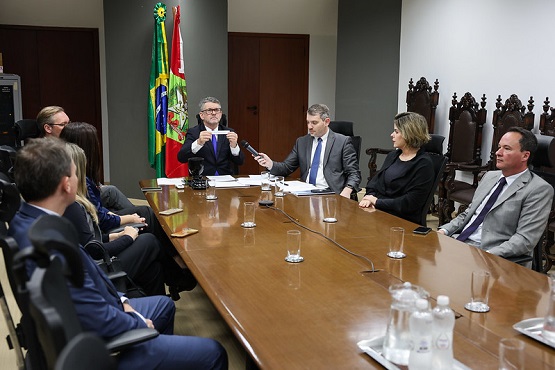 A imagem mostra uma reunião formal com várias pessoas sentadas ao redor de uma mesa de madeira. Há seis pessoas visíveis na foto, todas vestidas formalmente com ternos. No fundo, há duas bandeiras, uma do Brasil e outra de um estado ou município, que não é possível identificar claramente. A mesa está equipada com microfones e copos de água. A cena parece ser em um ambiente governamental ou corporativo, dado o estilo formal e a presença das bandeiras.
