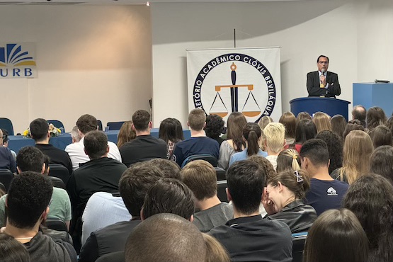 Foto horizontal: Francisco Oliveira Neto discursa em auditório da FURB, público atento, púlpito à direita, ambiente acadêmico, tons neutros.