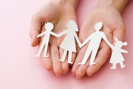 A imagem mostra duas mãos abertas segurando recortes de papel que formam a silhueta de uma família. Os recortes representam quatro figuras humanas: duas figuras maiores, que parecem ser adultos, e duas figuras menores, que parecem ser crianças. O fundo é de cor rosa.