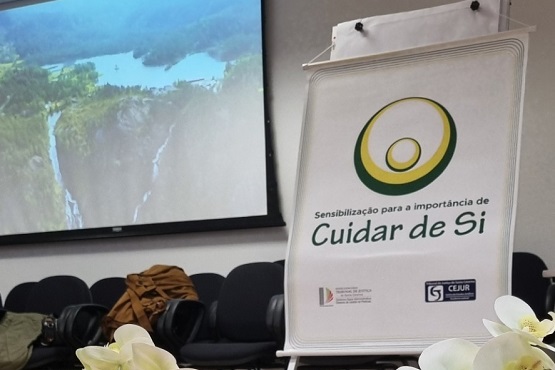A imagem mostra um banner com o texto "Sensibilização para a importância de Cuidar de Si". Há um logotipo ao lado do texto, que consiste em dois círculos concêntricos, um maior em verde e um menor em amarelo. Na parte inferior do banner, há o logotipo do "CRAS" e do "Governo de Santa Catarina". Ao fundo, há uma tela de projeção mostrando uma imagem de paisagem natural, possivelmente uma montanha ou colina com vegetação. À esquerda, há algumas cadeiras pretas e um arranjo de flores brancas.