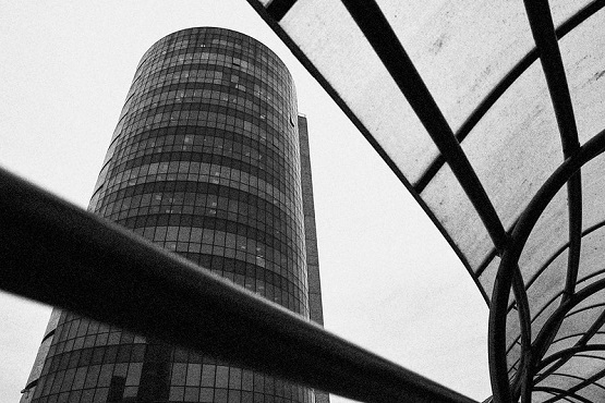 A imagem em preto e branco mostra um edifício alto e cilíndrico, com muitas janelas, que parece ser feito de vidro ou ter uma fachada espelhada. O prédio ocupa a parte esquerda da imagem. À direita, há uma estrutura curva que parece ser uma cobertura ou passarela, feita de metal e vidro, com linhas que criam um padrão geométrico. A perspectiva da foto é de baixo para cima, dando uma sensação de imponência ao edifício. O céu está nublado, contribuindo para o contraste da imagem.