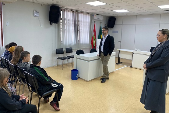 A imagem mostra uma sala de aula ou uma pequena sala de conferências. À esquerda, há um grupo de crianças sentadas em cadeiras, prestando atenção. Elas estão voltadas para um homem que está em pé, próximo a uma mesa branca no centro da sala. Ele está vestindo calça bege, camisa branca e um blazer preto. À direita, uma mulher está em pé, observando a cena. Ela está vestida com um casaco cinza escuro e óculos. No fundo da sala, há uma bandeira do Brasil e outra bandeira de Santa Catarina. As paredes são brancas e há janelas com persianas. O chão é de cor clara e o teto tem iluminação embutida.