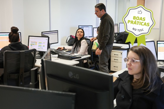 A imagem mostra um ambiente de escritório com várias pessoas trabalhando em computadores. Há quatro pessoas visíveis na foto. Três delas estão sentadas em frente a monitores, enquanto uma está em pé, aparentemente conversando com uma das pessoas sentadas. As mesas estão organizadas em um layout de escritório típico, com computadores e outros materiais de trabalho. No canto superior direito da imagem, há um selo verde com a inscrição "Boas Práticas no TJSC".