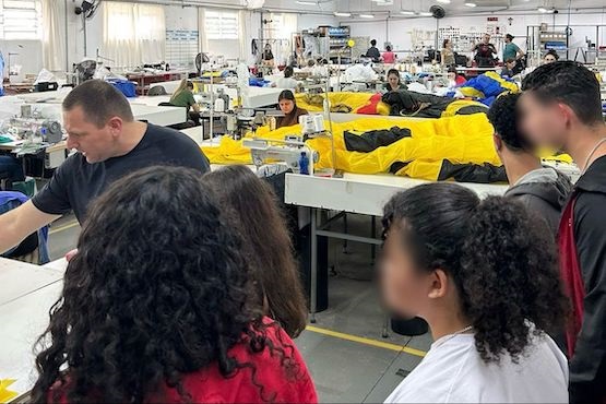 A imagem mostra o interior de uma fábrica ou oficina de costura. Há várias pessoas trabalhando em mesas grandes, onde estão espalhados tecidos amarelos e pretos, que parecem ser parte de roupas ou equipamentos. No primeiro plano, há um grupo de quatro pessoas, duas mulheres e dois homens, observando o ambiente. Um homem de camiseta preta está à esquerda, aparentemente explicando algo para o grupo. O local é bem iluminado, com várias máquinas de costura e ventiladores ao fundo. As pessoas estão concentradas em suas atividades, e o ambiente parece ser de trabalho colaborativo.