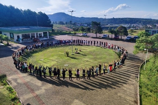 A imagem mostra um grande grupo de pessoas formando um círculo ao ar livre. Elas estão com os braços levantados, em torno de uma área gramada. Ao fundo, é possível ver uma construção com um telhado verde e algumas árvores. A paisagem ao redor inclui colinas e um céu azul com algumas nuvens. A cena parece ser de um evento ou atividade coletiva em um ambiente natural.