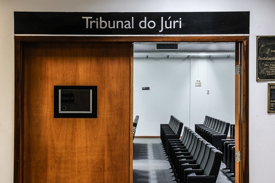 A imagem mostra a entrada de uma sala identificada como "Tribunal do Júri". A porta é de madeira e está parcialmente aberta, revelando o interior da sala. Dentro, há várias fileiras de cadeiras pretas organizadas em duas seções, todas voltadas para a frente da sala. As paredes internas são brancas e há iluminação no teto. Ao lado da porta, na parede, há uma placa de metal com inscrições, mas o texto não é legível na imagem.