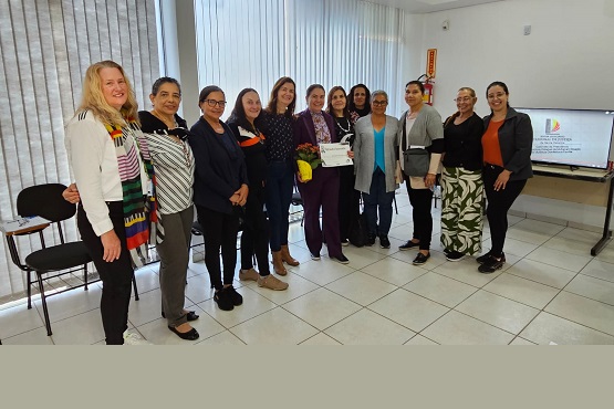 A imagem mostra um grupo de doze mulheres posando para uma foto em um ambiente interno. Elas estão em pé, lado a lado, e parecem estar em um tipo de evento ou reunião. Uma das mulheres, no centro, está segurando um certificado ou diploma. Todas estão sorrindo e parecem felizes. O ambiente é bem iluminado, com janelas ao fundo cobertas por persianas verticais. À direita, há uma televisão ou monitor que exibe um slide com a logo do TJSC. O chão é de azulejos claros e há algumas cadeiras vazias à esquerda. As mulheres estão vestidas de maneira casual e formal, com uma variedade de estilos e cores de roupas.