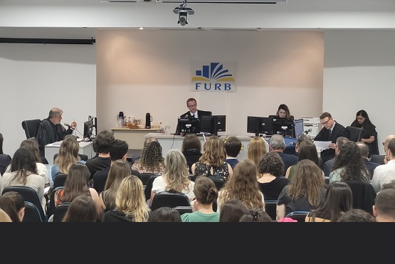 A imagem mostra o auditório da Universidade Regional de Blumenau (FURB), com várias pessoas sentadas, aparentemente assistindo a uma apresentação ou palestra. Na frente da sala, há uma mesa com cinco pessoas sentadas, duas delas usando computadores. No fundo, na parede, está o logotipo da FURB. A sala está cheia, com muitas pessoas sentadas em cadeiras, voltadas para a frente, onde ocorre a apresentação. A atmosfera parece ser formal, possivelmente um evento acadêmico ou uma sessão de julgamento simulado.