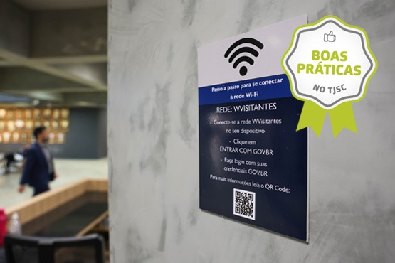 No topo do cartaz, há um ícone de sinal de Wi-Fi. As instruções dizem: "Passo a passo para se conectar à rede Wi-Fi". A rede mencionada é "WVISITANTES". As etapas incluem conectar-se à rede no dispositivo, clicar em "ENTRAR COM GOVBR" e fazer login com as credenciais GOV.BR. Há também um código QR na parte inferior do cartaz para mais informações. No canto superior direito da imagem, há um selo verde com a inscrição "BOAS PRÁTICAS NO TJSC". Ao fundo, é possível ver uma pessoa caminhando em um ambiente que parece ser um saguão ou área de espera, com móveis e uma parede com prateleiras ou expositores.