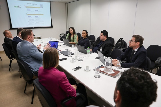 A imagem mostra uma reunião em uma sala de conferências. Há nove pessoas sentadas ao redor de uma mesa retangular branca. Na mesa, há laptops, copos, garrafas de água e alguns papéis. As pessoas estão vestidas formalmente, sugerindo um ambiente de trabalho ou reunião de negócios. Na parede ao fundo, há uma tela de projeção com uma apresentação exibida. A sala tem paredes brancas e parece bem iluminada.